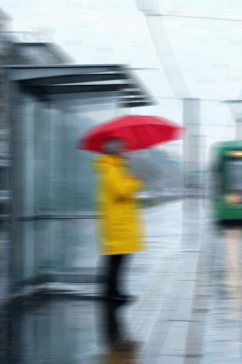 Photo of a woman in a yellow raincoat at a tram stop with heavy motion blur — before